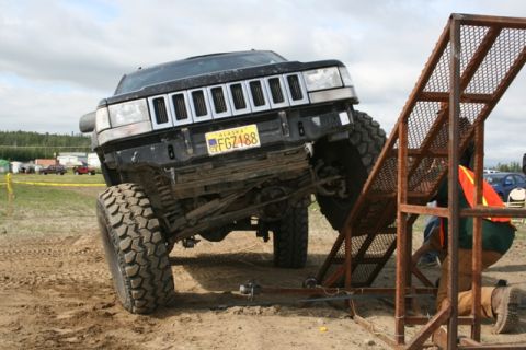 1990 Chevrolet Suburban - A lifted black Jeep Grand Cherokee with large off-road tires is positioned on a metal ramp during an off-road event. A person in an orange…