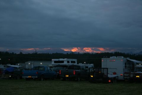 1990 Chevrolet Suburban - A fleet of pickup trucks and RVs are parked in a grassy field at dusk, with a mountain range illuminated by sunset in the background.