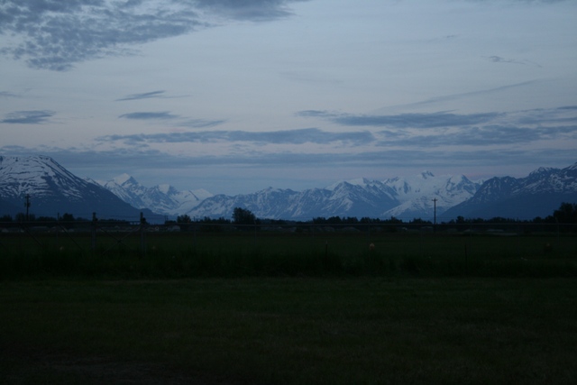 Snow-capped mountains under a cloudy sky, with a dark grassy field in the foreground.