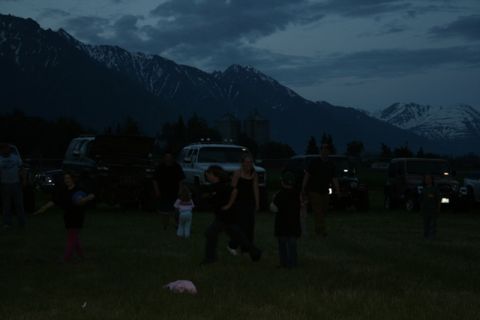 1990 Chevrolet Suburban - A group of people are gathered in a grassy field at dusk with a mountain range in the background. Several vehicles, including a dark-colore…