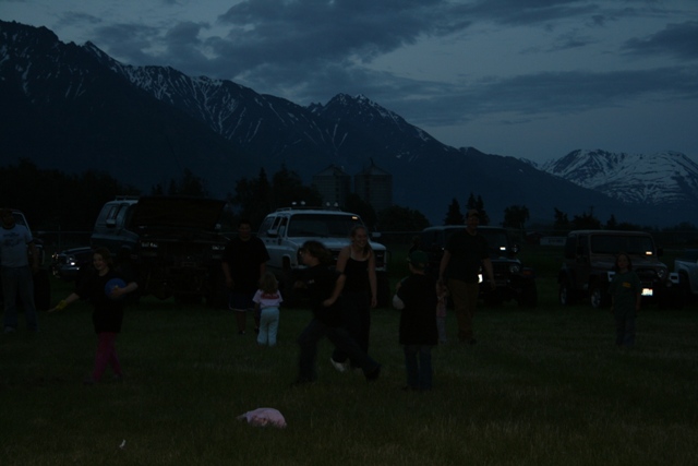 People gathered in a field at dusk with mountains and vehicles in the background.