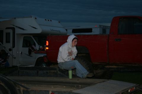 1990 Chevrolet Suburban - A person wearing a hooded sweatshirt and jeans sits on the edge of a trailer in front of a red pickup truck and a white RV. The sky is dark…