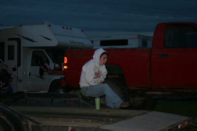 Person sitting on a trailer in front of a red pickup truck and white RV at dusk.