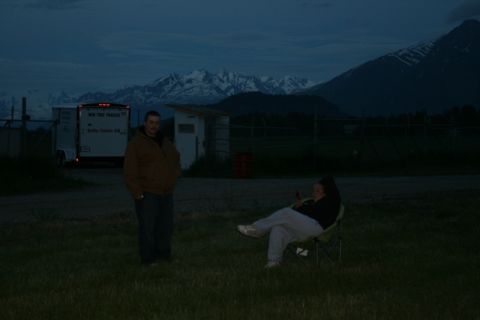 1990 Chevrolet Suburban - A man stands in a grassy field in front of a white trailer with text on the side, while a woman sits in a camping chair in the foreground.…