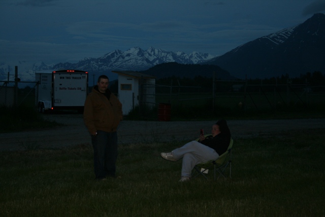 Man standing, woman sitting in chair, trailer, mountains, dusk.