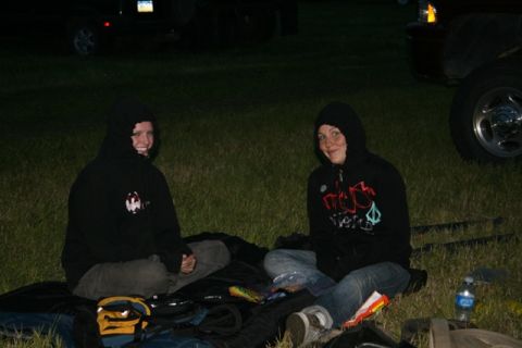 1990 Chevrolet Suburban - Two people in hooded sweatshirts sit on the grass in front of a dark-colored vehicle, possibly a Chevrolet Suburban, at dusk. The vehicle's…