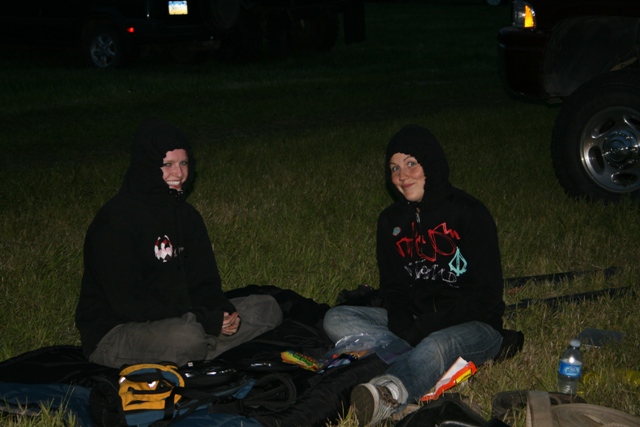 Two people sitting on grass at dusk in front of a dark SUV, with the vehicle's wheel visible.