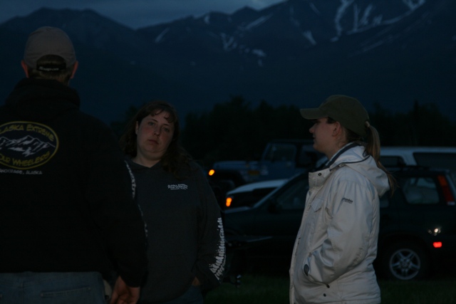 Three people stand in front of parked vehicles with mountains in the background.