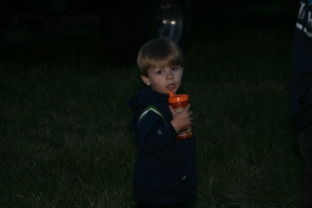 Young boy in a dark hoodie holding an orange sippy cup, standing in grass at dusk.