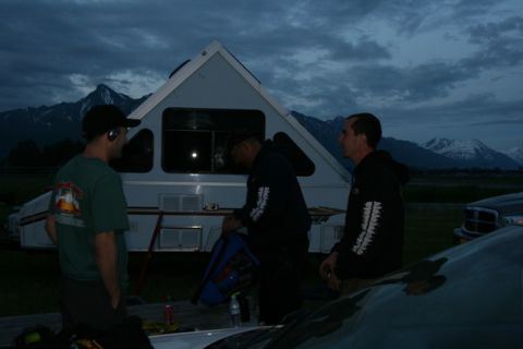 1990 Chevrolet Suburban - Three men stand near a pop-up camper trailer with mountains in the background during twilight. One man in a green t-shirt faces away from t…