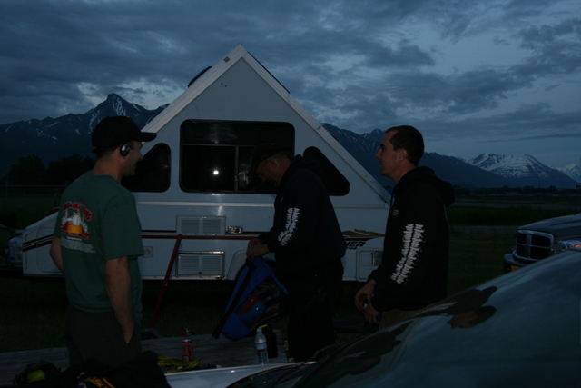 Three men by a pop-up camper trailer with mountains in the background at dusk.