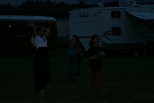 Four children holding balls in a field at dusk with a trailer and bus in the background.