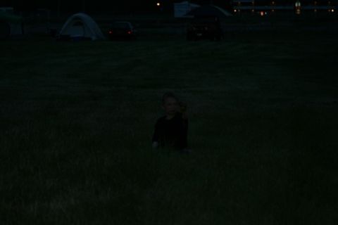 1990 Chevrolet Suburban - A young child sits in a grassy field at dusk, holding something in their right hand. In the background, a tent and two cars are visible.