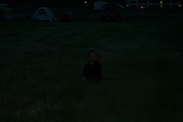 A child sits in a field at dusk with tents and cars in the background.