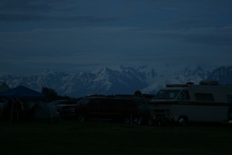 1990 Chevrolet Suburban - A dark, snow-capped mountain range is visible in the background behind a row of parked vehicles and tents at dusk. A large, dark SUV is par…