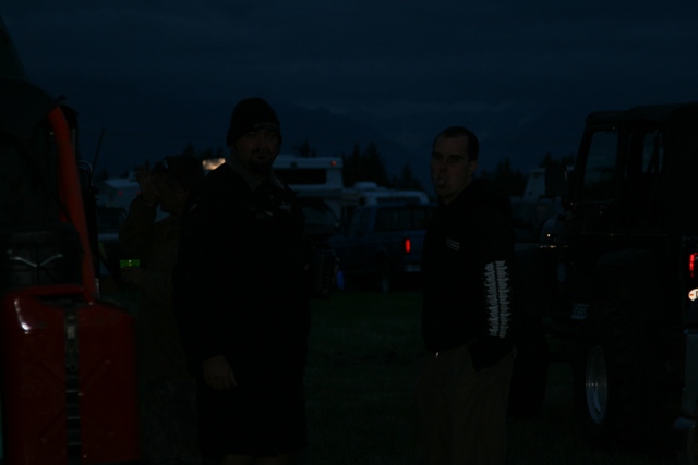 Two men stand in front of a dark SUV at dusk, with a red jerry can on the left.