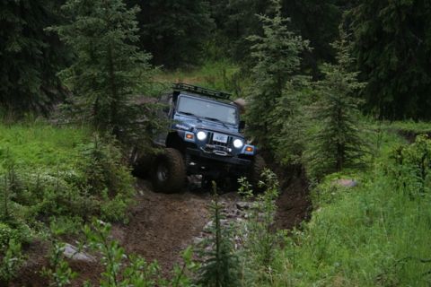 1990 Chevrolet Suburban - A dark blue Jeep Wrangler is navigating a muddy, rutted trail through a dense forest. The vehicle is angled downhill, with its tires deep i…