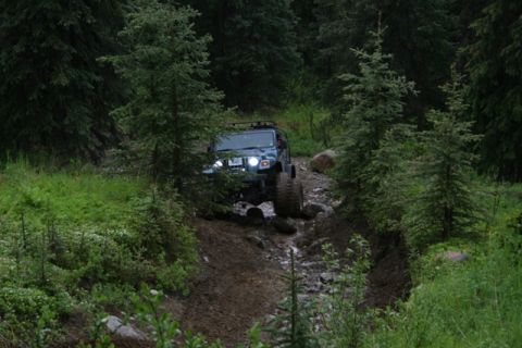 1990 Chevrolet Suburban - A dark blue Jeep Wrangler is navigating a muddy, rocky trail through a dense forest. The vehicle is angled slightly towards the viewer, wit…