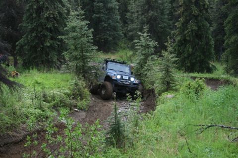 1990 Chevrolet Suburban - A blue Jeep Wrangler is navigating a muddy, rutted trail through a dense forest. The vehicle is angled downwards, with its front tires on a…