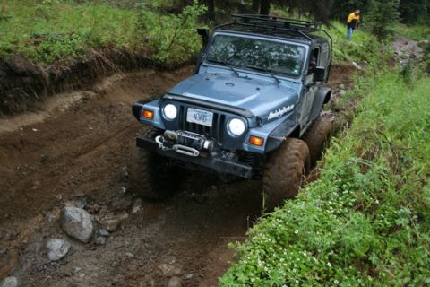 1990 Chevrolet Suburban - A blue Jeep Wrangler is navigating a muddy, rutted off-road trail with dense green foliage on either side. A person in a yellow jacket is v…