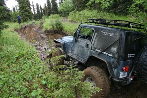1990 Chevrolet Suburban - A dark blue Jeep Wrangler is navigating a steep, muddy, and rutted off-road trail surrounded by dense green foliage and tall evergreen tree…