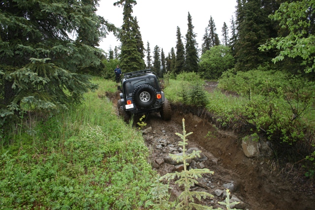 Dark Jeep Wrangler driving on a steep, muddy, and rocky off-road trail in a forest.