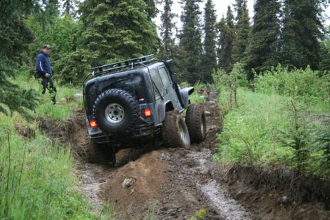 1990 Chevrolet Suburban - A black Jeep Wrangler is navigating a muddy, rutted trail surrounded by dense forest. A person stands on the side of the trail watching the…