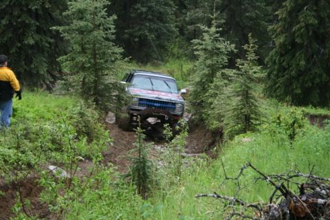 1990 Chevrolet Suburban - A dark colored Chevrolet Suburban is navigating a muddy trail through a dense forest. A person wearing a yellow and black jacket stands to…