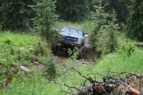 1990 Chevrolet Suburban - A lifted, older model Chevrolet pickup truck navigates a muddy, off-road trail surrounded by dense evergreen trees. The truck is angled tow…