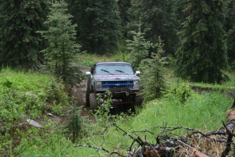 1990 Chevrolet Suburban - A lifted, older model Chevrolet pickup truck with a blue grille is driving off-road through a muddy, wooded trail. The truck has large tire…