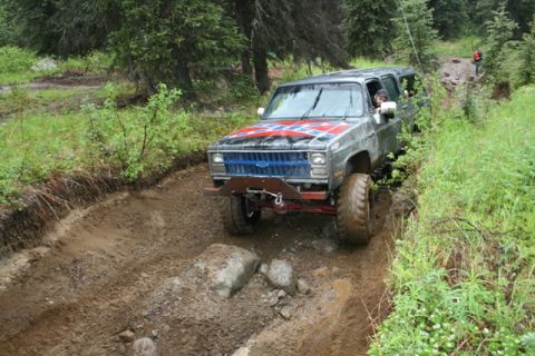 1990 Chevrolet Suburban - A lifted, dark gray Chevrolet Suburban with a Confederate flag decal on the hood navigates a muddy, rutted off-road trail in a wooded area.…