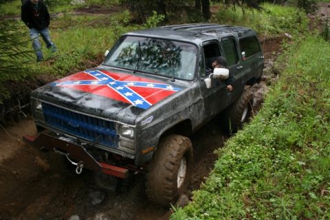 1990 Chevrolet Suburban - A dark gray Chevrolet Suburban with a Confederate flag decal on the hood is navigating a muddy, off-road trail. The vehicle is tilted and i…