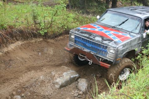 1990 Chevrolet Suburban - A gray Chevrolet Suburban with a Confederate flag decal on the hood navigates a muddy off-road trail with large rocks.