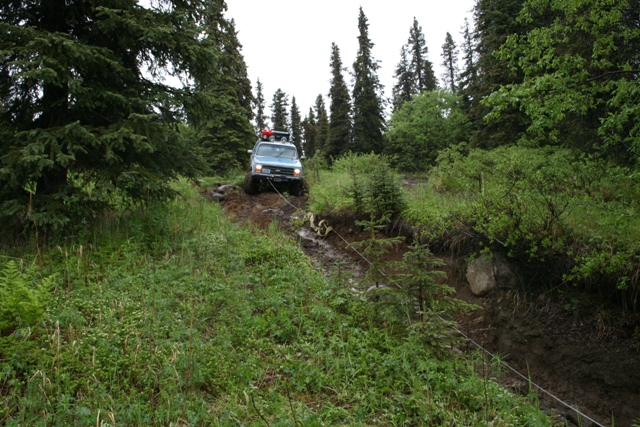 Light blue Chevrolet Suburban being winched up a muddy trail in a forest.