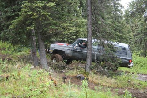 1990 Chevrolet Suburban - A gray Chevrolet Suburban is navigating a muddy, wooded trail, with one front wheel lifted off the ground. The driver is visible behind the…