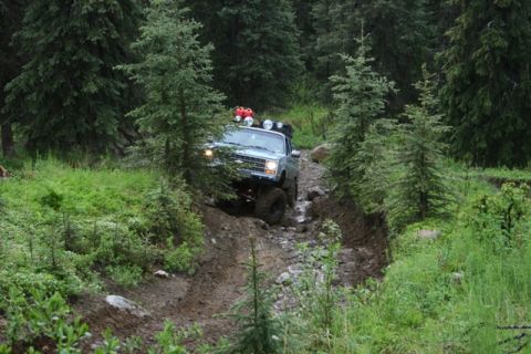 1990 Chevrolet Suburban - A light blue Chevrolet Suburban is driving down a muddy and rocky trail in a forest. The vehicle has a roof rack with red gas cans and auxi…