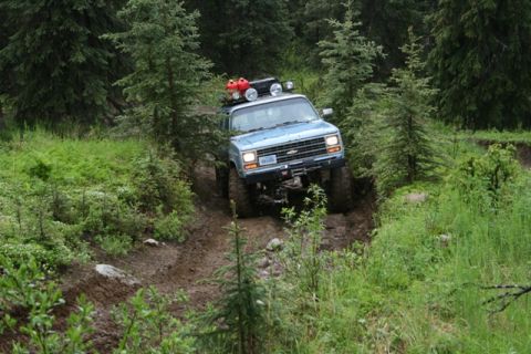 1990 Chevrolet Suburban - A light blue Chevrolet Suburban is navigating a muddy, off-road trail through a dense forest. The vehicle has a roof rack with two red gas…