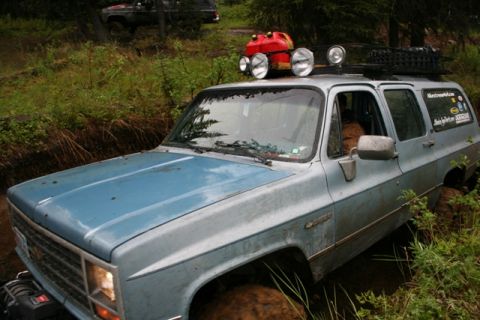 1990 Chevrolet Suburban - A light blue Chevrolet Suburban is navigating a muddy off-road trail, with a roof rack carrying a red jerrycan and spotlights. The vehicle…