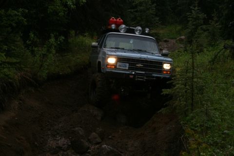 1990 Chevrolet Suburban - A lifted, blue Chevrolet SUV is navigating a steep, muddy trail in a dense forest. The vehicle has auxiliary lights and two red gas cans mo…