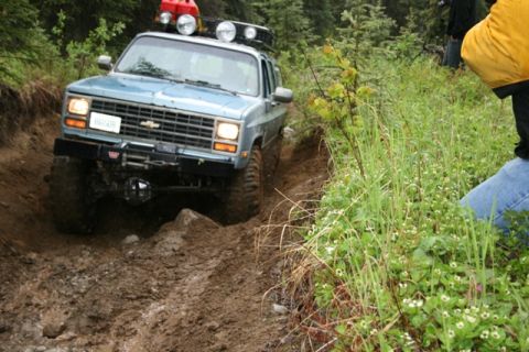 1990 Chevrolet Suburban - A light blue Chevrolet Suburban is navigating a muddy, rutted off-road trail surrounded by green foliage and trees. The vehicle has a roof…
