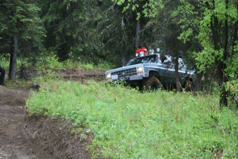 1990 Chevrolet Suburban - A light blue Chevrolet Suburban is driving off-road on a muddy track through a dense forest. The vehicle has off-road lights and a red stor…