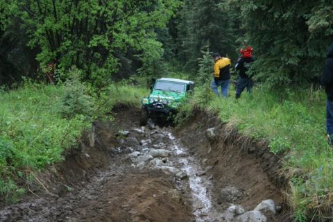 1990 Chevrolet Suburban - A green Jeep Wrangler navigates a muddy and rocky off-road trail surrounded by dense forest. Several people watch from the side of the trai…