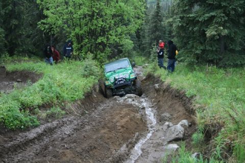 1990 Chevrolet Suburban - A green Jeep Wrangler navigates a muddy, rutted off-road trail with several people watching from the side.