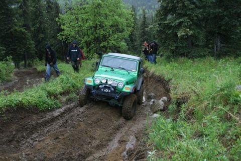 1990 Chevrolet Suburban - A bright green Jeep Wrangler is navigating a muddy off-road trail with several people watching and walking nearby in a wooded area.