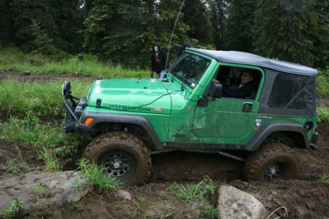 1990 Chevrolet Suburban - A green Jeep Wrangler Rubicon is stuck in thick mud on an off-road trail, with two people standing behind it and one person driving.