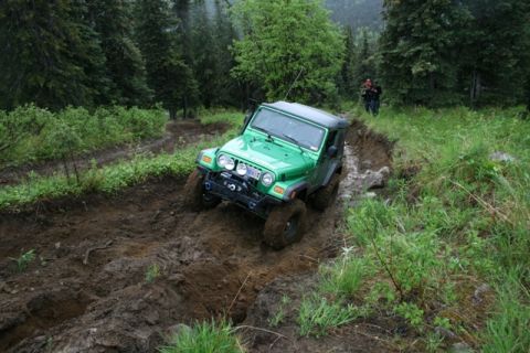 1990 Chevrolet Suburban - A green Jeep Wrangler is navigating a muddy, rutted trail through a dense forest. Two people are visible in the background on the side of t…