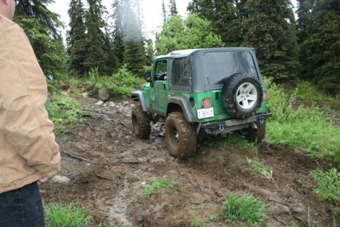 1990 Chevrolet Suburban - A green Jeep Wrangler is navigating a muddy, wooded trail. The vehicle is viewed from the rear-right, showing its off-road tires and spare…