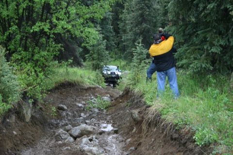 1990 Chevrolet Suburban - A white SUV navigates a muddy, rocky trail through a dense forest. Two people are standing on the side of the trail, watching the vehicle.