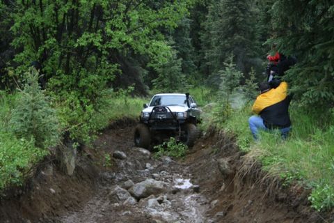 1990 Chevrolet Suburban - A white Toyota pickup truck with large tires navigates a muddy, rocky trail through a dense forest. Two people are visible on the right sid…