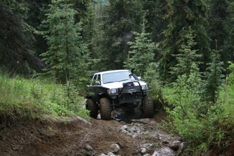 1990 Chevrolet Suburban - A white SUV with off-road modifications navigates a muddy and rocky trail through a dense forest.